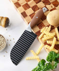 Side view of crinkle cutter knife with brown rubber handle and ribbed black blade on cutting board beside potato fries