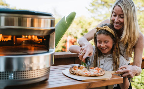 Mother and daughter using Pizza Cutter Wheel With Durable 304 Stainless Steel Blade And Powder Coated Aluminum Handle Dishwasher Safe Easy Grip For Quick Slices Perfect For Sharing With Family And The Crew Pi Collection to slice pizza outdoors.