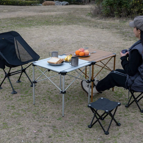 Ultra Lightweight Stable Folding Aluminum Table With Roll Up Top And Carry Bag For Camping Picnics Beach Chill Backyard Barbecues Tailgating And RV Trips Anywhere set up outdoors with chairs and food.