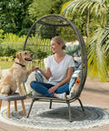 Woman sitting in patio hanging egg chair with black cushion in garden setting next to a dog on a stool.