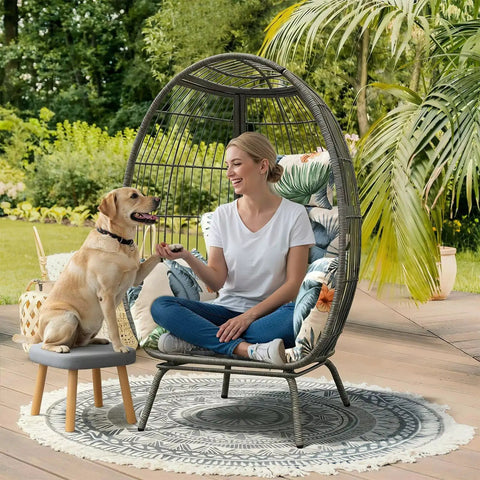 Woman sitting in patio hanging egg chair with black cushion in garden setting next to a dog on a stool.