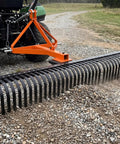 Heavy Duty 6 Foot Tractor Landscape Rake With 37 Steel Tines, 360 Degree Rotation, 3 Point Hitch in use on gravel path, orange