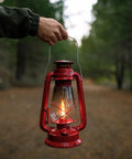 12 Inch Hurricane Lantern With Durable Red Finish For Camping And Emergencies held outdoors on a forest path.