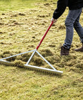 Man using all aluminum landscape rake with 66 inch handle and 36 inch wide head for lawn care, loosening soil on grassy area.