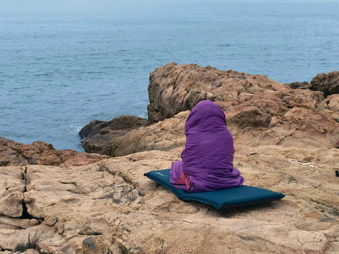 Person in an Ultralight Camping Sleeping Bag For Adults And Kids on rocky coastline, focused on sleeping bag features.