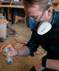 Person wearing Pro Grade Half Mask Respirator With Reusable Filters And Safety Glasses For Dust Pollen And Organic Vapor Protection Includes Cartridges And Extra Filters spraying aerosol in workshop