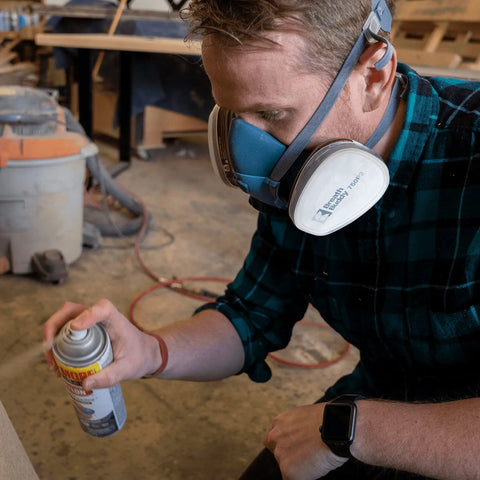 Person wearing Pro Grade Half Mask Respirator With Reusable Filters And Safety Glasses For Dust Pollen And Organic Vapor Protection Includes Cartridges And Extra Filters spraying aerosol in workshop