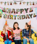 Three children celebrating a birthday with a cake and balloons, wearing party hats under a happy birthday banner.