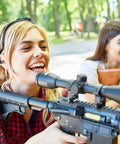 Two women outdoors, one holding a scoped rifle, both smiling, with trees in the background.