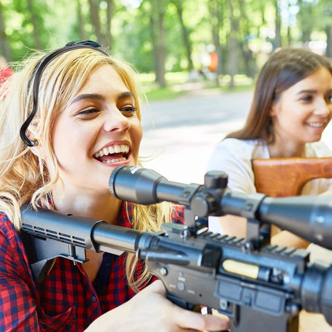 Two women outdoors, one holding a scoped rifle, both smiling, with trees in the background.