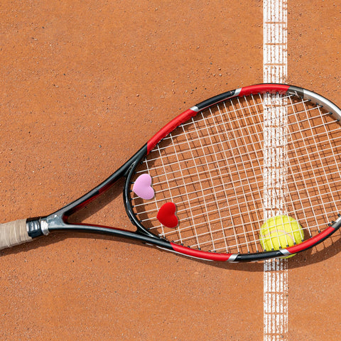 Heart Shaped Tennis Dampeners Set Of 4 Silicone Vibration Absorbers in red and pink on a racket near a tennis ball on clay court