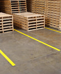 Heavy Duty Polyester Floor Marking Tape In Bright Yellow With Kick Ass Adhesion For Concrete Warehouses and 2 Inch By 100 Foot Roll on warehouse floor beside wooden pallets.