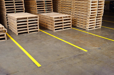 Heavy Duty Polyester Floor Marking Tape In Bright Yellow With Kick Ass Adhesion For Concrete Warehouses and 2 Inch By 100 Foot Roll on warehouse floor beside wooden pallets.