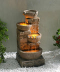 Outdoor Water Fountain With LED Lights, Cascading Bowls And Stacked Stone Look, freestanding on white stones, surrounded by plants.