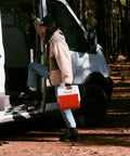 Person holding Playmate Portable Cooler With Tent Top Design, red, near van in forest, ideal for camping and road trips.