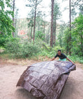 Person setting up Rip Stop Tarp 8 By 10 Feet Heavy Duty Waterproof Brown Outdoor Shelter For Camping And Backyard Use in a forest