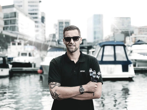 Man standing by marina holding a product, wearing sunglasses and black polo shirt, boats in the background.