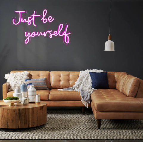 Living room with pink USB powered neon sign 'Just be yourself' on wall, above a tan leather sofa and wooden coffee table.