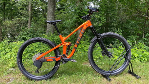 Orange mountain bike with Rear Derailleur Guard For Mountain Bikes And EBikes, Ultra Lightweight And Easy To Install, Universal Fit No Modifications Needed, Protect Your Derailleur From Rocks, Roots And Obstacles On The Trail Made In USA in a forest setting.