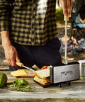 Person using XL Cast Iron Pie Iron Sandwich Maker For Campfire Cooking on wooden table with ingredients nearby