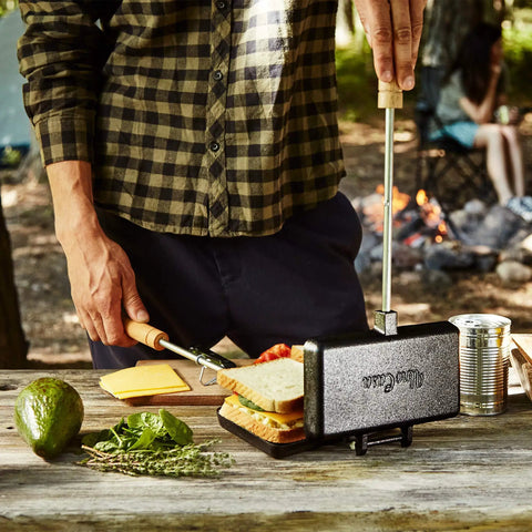 Person using XL Cast Iron Pie Iron Sandwich Maker For Campfire Cooking on wooden table with ingredients nearby