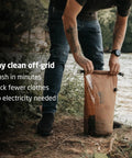 Compact Travel Laundry Washbag Featuring Patented Flex Washboard Design For Machine Grade Dirt Removal Without The Laundromat Hustle On Road Trips Camping Adventures Or Hotel Hops, being used by a camper to wash clothes by a river.