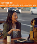 Woman at outdoor table with a coffee, backpack, and notebooks, discussing social connectivity; cityscape in background.