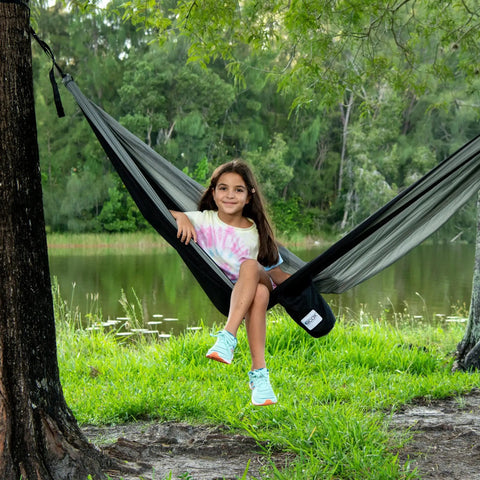 Girl sitting in Parachute Nylon Hammock Single With 2 Tree Straps by a lake, ultra portable lightweight camp gear