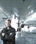 Aircraft engineer standing confidently under a large jet in a hangar, wearing a black jacket with arms crossed.