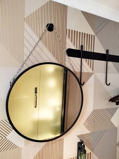 Round mirror hanging on chain against geometric patterned wall in well-lit room with hooks nearby.