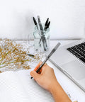 Handwriting in a notebook with BIC Round Stic Xtra Life black ballpoint pens, surrounded by a laptop and dried flowers.