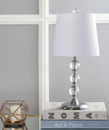 Clear Crystal 20 Inch Mini LED Table Lamp In Chrome Finish With White Linen Drum Shade on books against paneled wall.