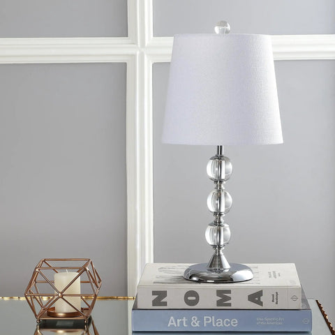 Clear Crystal 20 Inch Mini LED Table Lamp In Chrome Finish With White Linen Drum Shade on books against paneled wall.