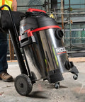 16-Gallon Stainless Steel Wet Dry Shop Vacuum With Cart in use on a construction site, showing large rear wheels and push handle.