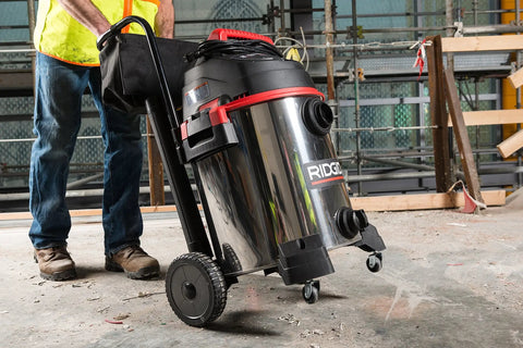 16-Gallon Stainless Steel Wet Dry Shop Vacuum With Cart in use on a construction site, showing large rear wheels and push handle.