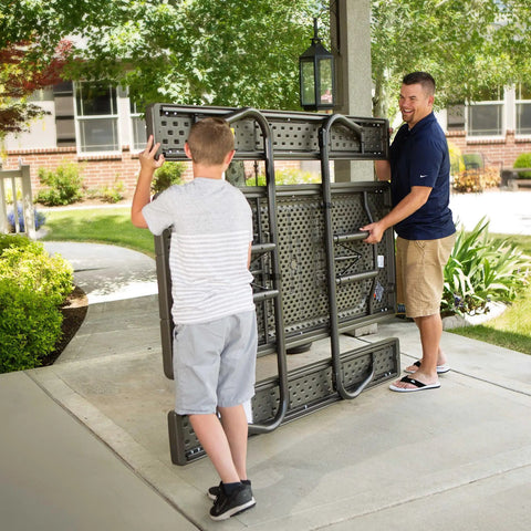 Two people folding a Six Foot Folding Picnic Table With Classic Rugged Steel Frame Seats Eight UV Protected Top Weather And Rust Resistant Flat Fold For Easy Stow And An Umbrella Hole For Backyard Hangouts.