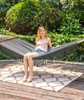 Person relaxing on Cotton Double Hammock With 55 Inch Spreader Bar, Portable Camping Hammock Kit, Dark Gray, in garden setting.