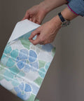 Close-up of hands holding Self Adhesive Privacy Glass Window Film 17 Inch By 78 Inch Roll Stained Glass Blue Green with gridlines.