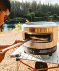 Man cooking pizza outdoors with Outdoor Pizza Oven On The Go For Gas Or Propane Cooking, Portable Cordierite Stone Surface.