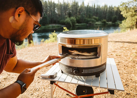 Man cooking pizza outdoors with Outdoor Pizza Oven On The Go For Gas Or Propane Cooking, Portable Cordierite Stone Surface.