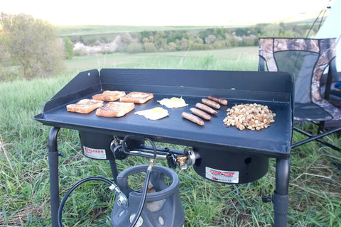 Deluxe Double Burner Steel Griddle With Extra Large 14 Inch By 32 Inch Surface Even Heat Diffusion From Built In Diffuser Plates Pre Seasoned Steel Finish For Easy Outdoor Camping And Backyard Cooking set up outdoors with food cooking on it