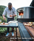 Outdoor Pizza Oven On The Go For Gas Or Propane Cooking, Portable Cordierite Stone Surface, Demi Dome Design And Wide Mouth Opening, Stainless Steel Build, Quick Preheat And Piping Hot Pizza Ready In Under Two Minutes on a picnic table with a man preparing pizza next to it.