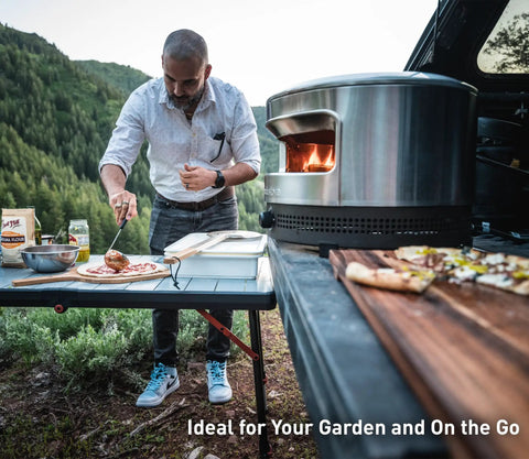 Outdoor Pizza Oven On The Go For Gas Or Propane Cooking, Portable Cordierite Stone Surface, Demi Dome Design And Wide Mouth Opening, Stainless Steel Build, Quick Preheat And Piping Hot Pizza Ready In Under Two Minutes on a picnic table with a man preparing pizza next to it.