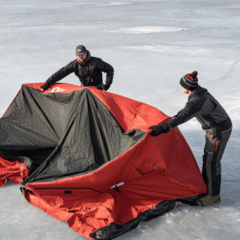 Two men setting up Quickfish Six Person Portable Pop Up Ice Fishing Shelter With Fully Quilted Insulation No Trip Door And Deluxe Anchors For Hell Of A Winter Setup In Red Black on ice.