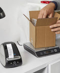 Turbo Driven Direct Thermal Label Printer With Automatic Size Detection And Plug And Play Usb Hookup Plus Lan Networking For Offices Or Shipping Without Any Ink Or Toner Hassle Just A Smooth Operator Ready To Roll Out The Daily Grind next to a cardboard box being filled.