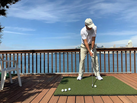 Golfer using Beefy 5x5 Golf Practice Mat With Realistic Fairway Feel Nylon Surface Indoor Outdoor Turf on a wooden deck by the water.