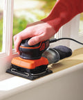 Quarter Sheet Orbital Palm Sander Professional Grade 220 Watt Motor Featuring A Snappy Pressure Trigger Switch Lightweight Orange Body in use on window frame.