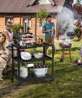 Outdoor scene with Grill Cart Table That Rolls On Wheels For Indoor And Outdoor Use with kitchenware, drinks, and pizza oven.