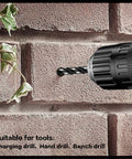 Side view of a black cordless drill with a masonry bit drilling into a brick wall; plant leaves visible left