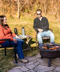 People sitting by Bronze 30 Inch Outdoor Wood Burning Fire Pit With Grill Grate, showing sausages cooking on a sunny day.